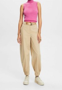 Nainen, joka on pukeutunut hihattomaan vaaleanpunaiseen ribattuun crop-topiin, vaaleisiin korkeavyötäröisiin housuihin, joissa on vyö, ja vaaleisiin tennareihin, joissa on nilkkasukat.