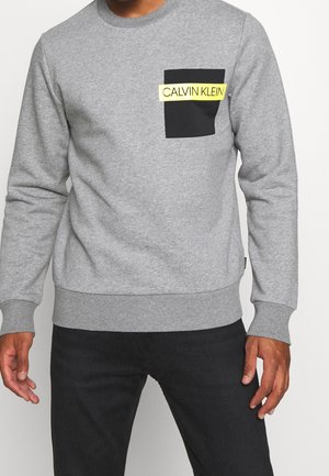 Mann trägt graues Sweatshirt mit langen Ärmeln, schwarzer Brusttasche und gelbem Streifen mit der Aufschrift „CALVIN KLEIN“, kombiniert mit einer schwarzen Hose.