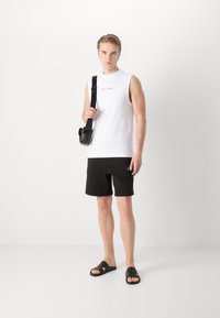 Camisa sin mangas blanca con el texto "Esto es amor", pantalones cortos negros y sandalias negras. El modelo sostiene un bolso cruzado negro. Fondo simple.