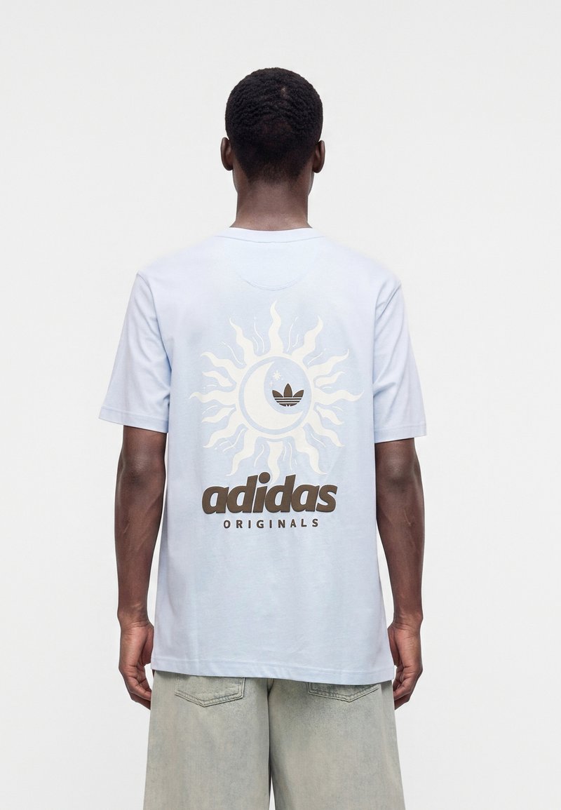 Άνδρας που φοράει ανοιχτά μπλε t-shirt Adidas Originals με λογότυπο ήλιου και φεγγαριού στην πλάτη, σε συνδυασμό με ανοιχτό γκρι σορτς, γυρισμένος με την πλάτη.