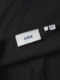 Svart stoff med en glatt tekstur; etiketten har merkevaren "JJXX" i blått på hvit bakgrunn, størrelse "S," og "LAGET I BANGLADESH."