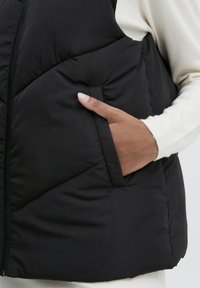 Gilet en duvet noir avec fermeture zippée, présentant un design matelassé diagonal et une poche latérale. Le tissu est lisse avec une finition mate.