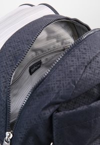 Granatowy plecak z zewnętrzną powierzchnią w heksagonalny wzór. Wnętrze z paskowanym podszewką i zamykaną na zamek kieszenią. Widoczna metka marki.