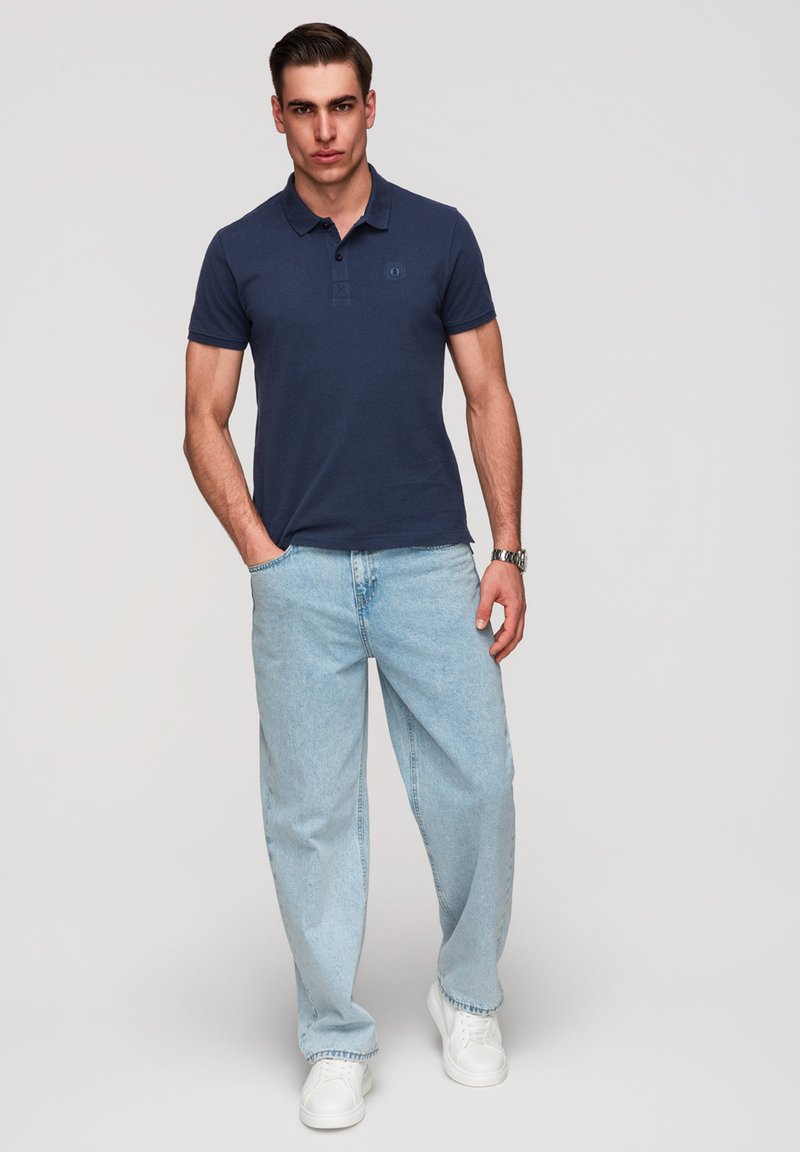 Jeune homme portant un polo bleu marine, un jean clair ample, des baskets blanches et une montre-bracelet, debout avec une main dans la poche.