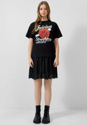 Junge Frau trägt ein schwarzes "Juiciest Strawberry"-Grafik-Shirt, schwarzen Plisseerock und schwarze Schnürstiefeletten, steht vor einem schlichten Hintergrund.