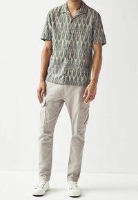 Man in een korte grijze gemuste overhemd met knoopjes, gecombineerd met lichtbeige cargobroeken en witte sneakers tegen een effen achtergrond.