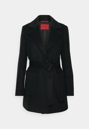 Cappotto in lana nera con colletto a revers, vita cinturata e due tasche frontali; linee pulite e texture liscia, con un'etichetta rossa all'interno.