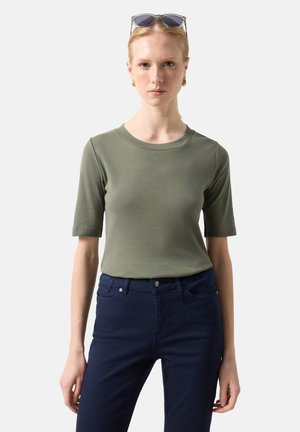 Ung kvinde iført olivengrøn kortærmet top, mørkeblå jeans, guldøreringe i hoops og solbriller, der hviler på hendes hoved mod en ensfarvet baggrund.