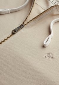 Sudadera cremosa con cremallera fabricada en tejido texturizado, con una cremallera dorada, cordones blancos y un pequeño logo bordado en el pecho.