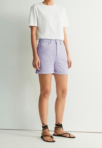Fialové bavlněné šortky s ohrnutým lemem, předními kapsami a zapínáním na knoflík, v kombinaci s jednoduchým bílým tričkem s kulatým výstřihem a černými sandály na řemínky.