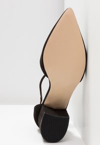 Chaussure slingback noire en daim à bout pointu, inclinée contre un mur blanc, montrant une semelle beige texturée et un petit talon carré.