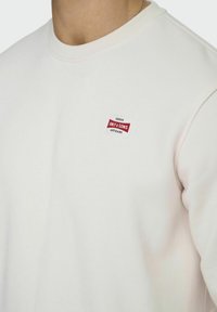 Licht beige sweatshirt met een ronde hals, getextureerde katoenstof en een rood-wit logopatch op de borst.