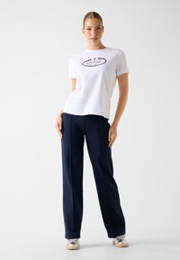 Weißes Baumwoll-T-Shirt mit schwarzem "GUESS"-Logo, kombiniert mit navyblauen, hochtaillierten Hosen und silbernen Sneakers, vor hellem Hintergrund stehend.