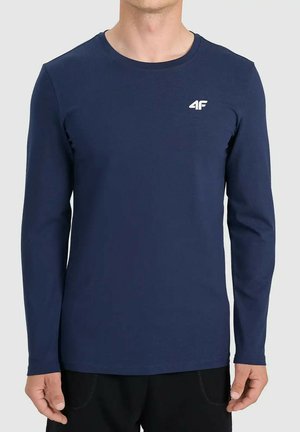 Mężczyzna w granatowej koszulce z długim rękawem z małym białym logo „4F” na lewej piersi, stojący na jednolitym tle.