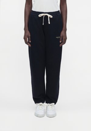 Personne portant un jogging noir Armani Exchange avec un cordon blanc et des baskets blanches, debout devant un fond uni.