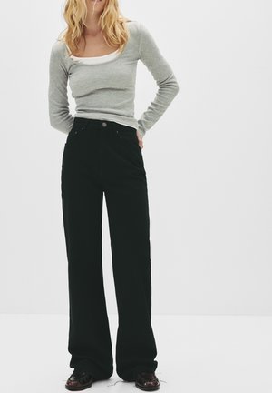 Vrouw met blond haar die een grijs shirt met lange mouwen, zwarte broek met wijde pijpen en donkere schoenen draagt, staand tegen een witte achtergrond.
