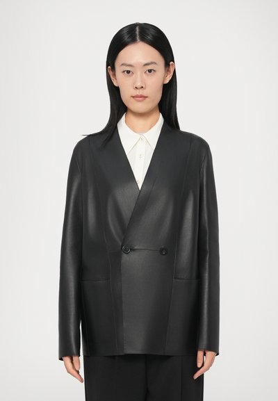 Blazer noir en cuir à double boutonnage avec deux poches frontales, texture lisse et boutons. Chemise blanche à col portée en dessous.