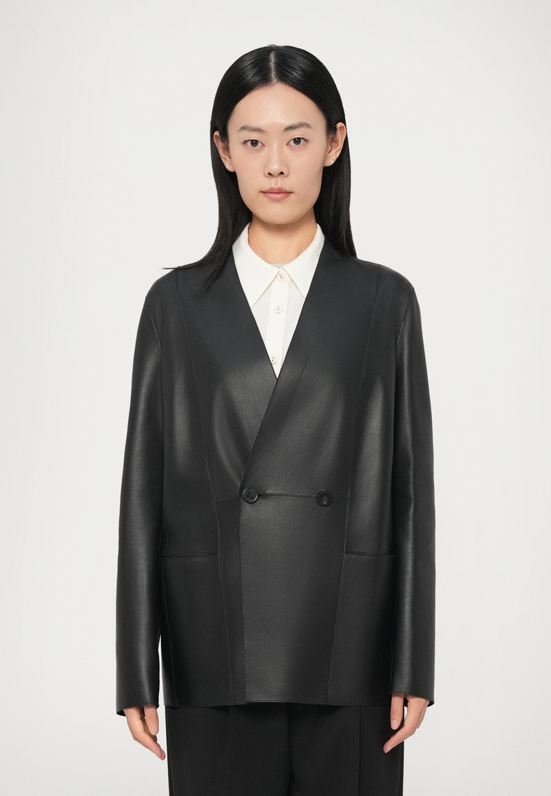 Melnā ādas dubultpogas žakete ar divām priekšējām kabatām, gluda tekstūra un pogas. Valkāta zem tā balta krekla ar apkakli.