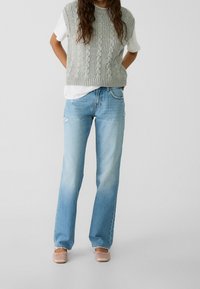 Chaleco de punto gris sobre una camiseta blanca, combinado con jeans claros rotos. Corte relajado y diseño de pierna ancha. Textura suave y estilo casual.