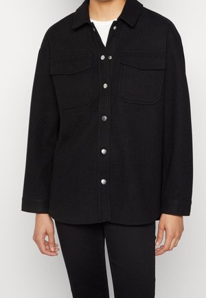 Personne portant une veste noire à boutons avec deux poches poitrine et des manches longues sur une chemise blanche.