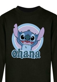 Melna sporta krekls ar karikatūras tēlu, kuram ir zilas spalvas un izteiktas ausis. "Ohana" teksts zem tēla uz gaiši zila apļa fona.