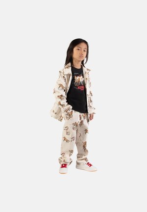 Enfant debout avec assurance, portant une veste crème et un pantalon ornés d'imprimés de chevaux, un t-shirt graphique noir et des baskets blanches aux lacets rouges.