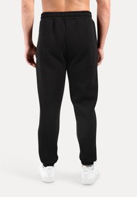 Muž stojící, oblečený v černých teplácích a bílých teniskách Nike Air, zobrazený zezadu na jednobarevném světlém pozadí.