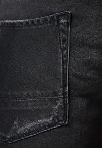 Tela de mezclilla negra con un bolsillo trasero que presenta costuras en contraste y un ligero desgarro en el borde, con una textura general suave.