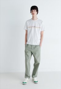 Fehér pamut póló színes logós csíkkal, világoszöld pamut nadrággal párosítva és zöld részletekkel ellátott fehér sportcipővel.