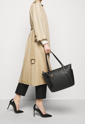 Persona con abrigo beige, pantalones negros recortados, tacones altos negros, sosteniendo un gran bolso de cuero negro con detalle de borla dorada.