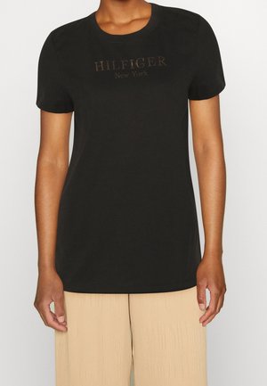 Persoon draagt een zwart T-shirt met korte mouwen met de tekst "HILFIGER New York" en beige geribbelde broek, staand tegen een effen witte achtergrond.