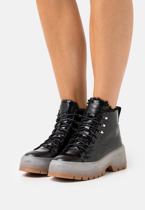 Bottines en cuir verni noir avec un col en fourrure, présentant des œillets argentés, des lacets noirs et une semelle en caoutchouc transparente avec des accents marron.