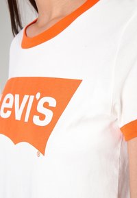 Lähikuva henkilöstä, joka pukeutuu valkoiseen T-paitaan, jossa on oranssi kaulus, hihansuut ja osittainen oranssi tekstilogo rinnassa.
