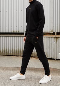 Zwarte langlevende top en broek van zachte stof, met een hoge kraag en vooraan branding. Gecombineerd met witte sneakers.