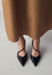 Czarne skórzane szpilki z ostrym czubkiem, z krzyżującymi się paskami i smukłym designem, zestawione z brązową, lejącą się spódnicą.