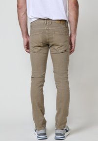 Beige slim fit farmernadrág öt zsebbel, részletes varrással és bőr folttal a deréknál. Szürke sportcipővel viselve.