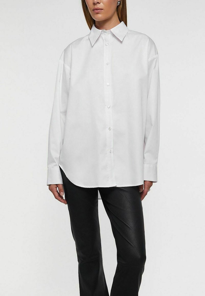 Valkoinen napitettava paita, jossa klassinen kaulus, pitkät hihat ja rento istuvuus, valmistettu sileästä kankaasta. Yhdistetty mustiin istuviin housuihin.