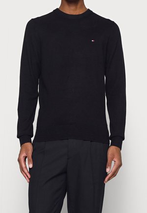 Mann trägt einen eng anliegenden schwarzen Langarmpullover mit Rundhalsausschnitt und einem kleinen rechteckigen Logo in Rot, Weiß und Blau auf der Brust.
