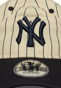 Beige baseballcap med marineblå striper, med en brodert marineblå NY-logo. Svart skjermbunn med justerbar stropp. New Era-merkelapp synlig.