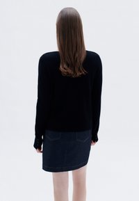 Schwarzer Pullover mit langen Ärmeln und Knopfdetails, kombiniert mit einem dunklen Jeansrock, der über zwei Gesäßtaschen und kontrastierenden Nähten verfügt.