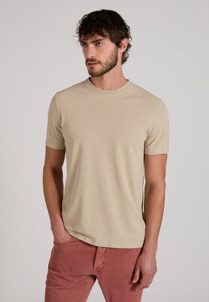 Uomo con capelli corti scuri e barba che indossa una t-shirt beige a maniche corte e pantaloni color ruggine, in piedi con una mano in tasca.