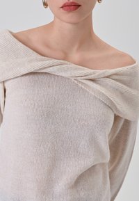 Maglione lavorato a maglia in beige chiaro con design scollo a spalla e dettagli del collo intrecciati. Tessuto morbido con un sottile motivo discreto.