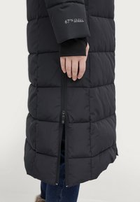 Sort pufferjakke med quiltet mønster, har en frontlynlås og tekstdetalje på ærmet. Stoffet ser glat og isoleret ud.