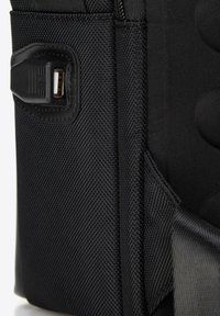 Sac à dos noir avec un tissu texturé, une fermeture éclair et un port USB sur le côté pour la connectivité des appareils électroniques.