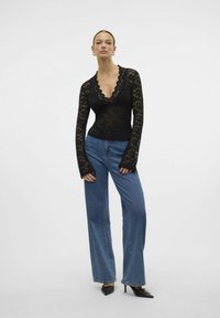 Top en dentelle noire avec décolleté festonné et manches longues, associé à un jean bleu taille haute évasé. La mannequin porte des chaussures à talons noires.