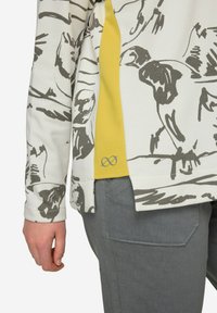 Chemise à manches longues blanche avec des illustrations d'oiseaux gris, se distinguant par un panneau latéral jaune. Portée avec un pantalon gris, présentant une coupe décontractée.
