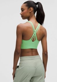 Zielony stanik sportowy z krzyżującymi się ramiączkami, o dopasowanym kroju i gładkiej teksturze. Noszony z jasnooliwkowymi spodniami sportowymi.