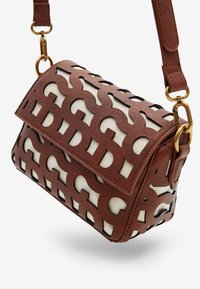 Brązowa skórzana torba na ramię z wyciętym geometrycznym wzorem, odsłaniającym białą tkaninę pod spodem, z elementami w kolorze złotym i regulowanym paskiem.
