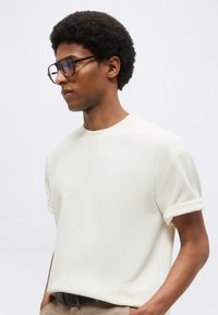 Άνδρας με κοντά σγουρά μαλλιά, φοράει μεγάλα γυαλιά, λευκό υφασμάτινο πουκάμισο με κοντά μανίκια διπλωμένα και μπεζ παντελόνι με μαύρη ζώνη.