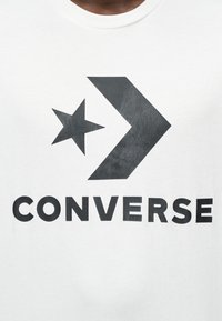 Άσπρο μπλουζάκι με μεγάλο μαύρο λογότυπο "CONVERSE" και γραφικό με αστέρι και κατευθυντικό βέλος από κάτω, φτιαγμένο από μαλακό βαμβακερό ύφασμα.